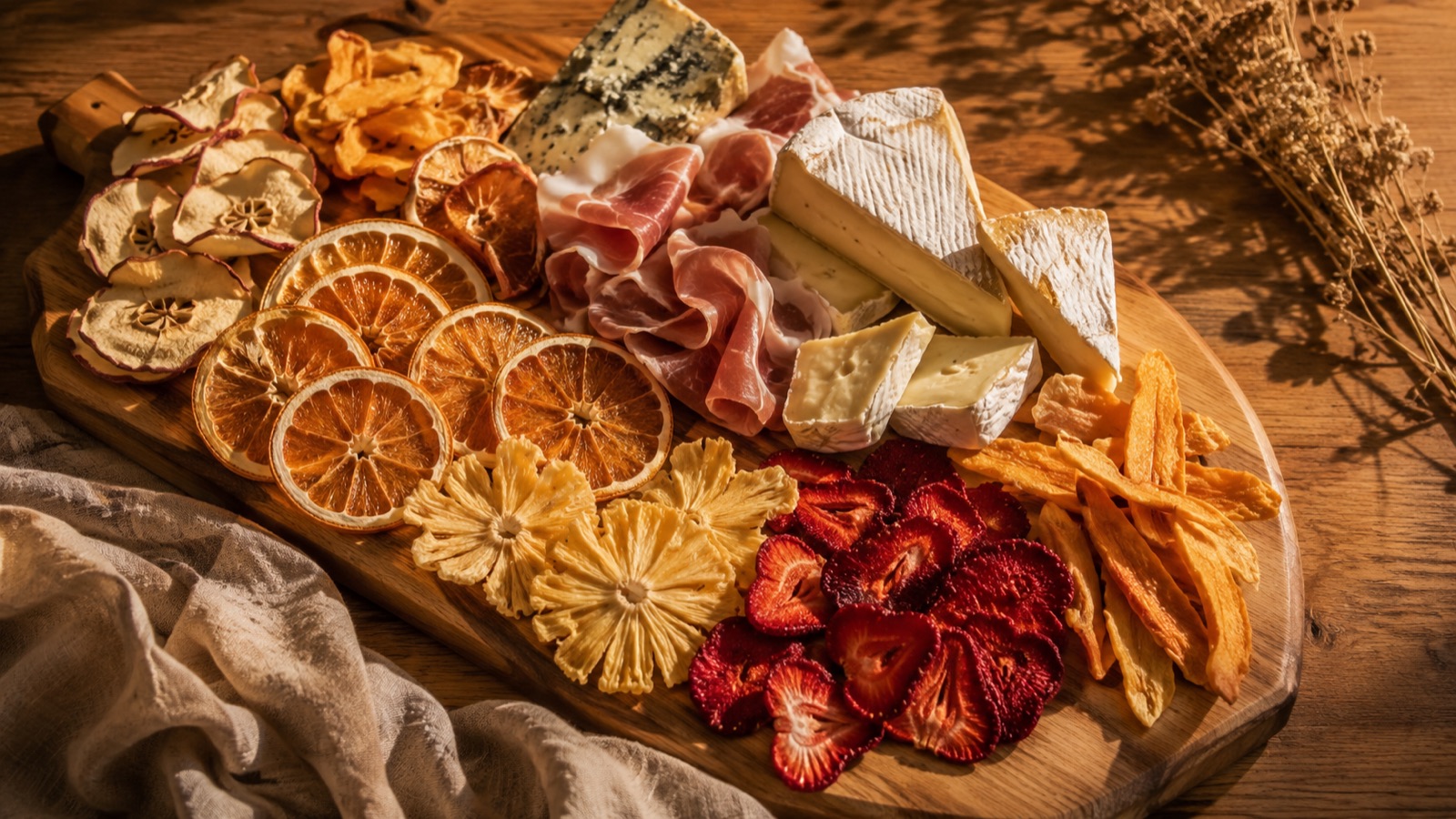 A wooden grazing board with cheese, cured meat, and clusters of dried citrus wheels and dried apple, mango, pineapple, and strawberry